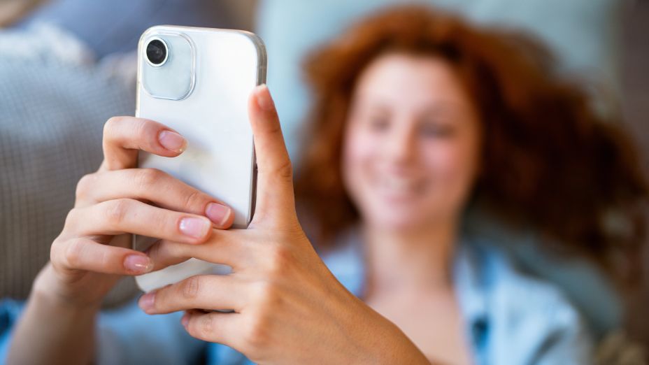 Im Vordergrund sieht man zwei Hände, welche ein Handy halten. Im Hintergrund sieht man verschwommen eine Frau.