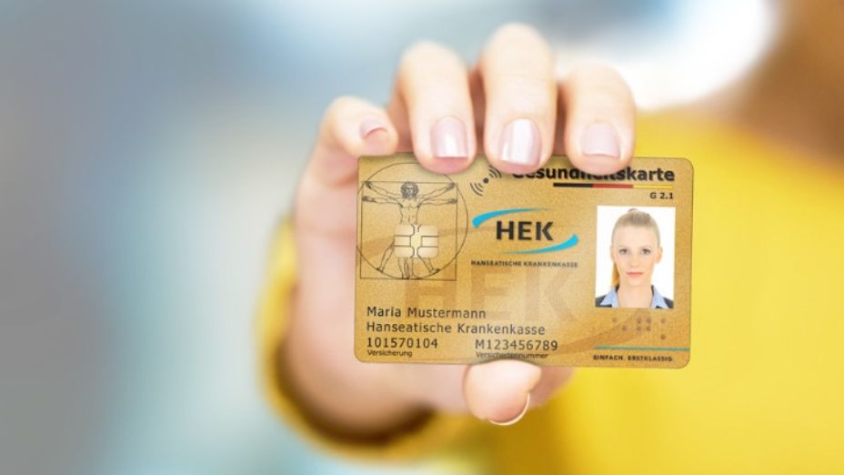 Im Vordergrund ist die goldene Versichertenkarte der HEK zu sehen. Verschwommen dahinter steht eine Person in einem Oberteil, welches gelb ist.