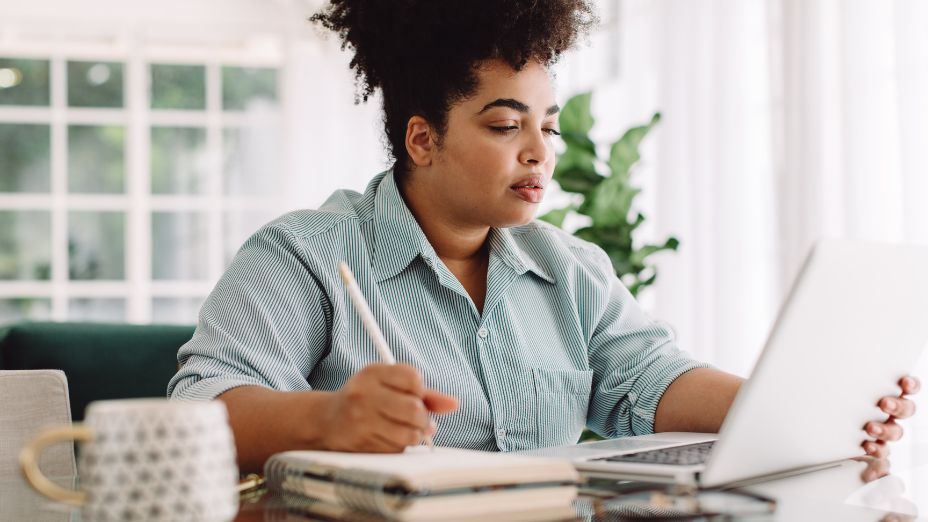 Eine Frau sitzt am Tisch, hält mit der linken Hand ihren Laptop fest und schreibt mit der rechten Hand auf einem Block.