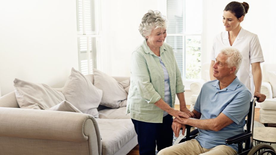 Ein Mann sitzt im Rollstuhl und hält die Hand seiner Frau, welche neben ihm steht. Hinter dem Mann im Rollstuhl steht die Pflegerin.