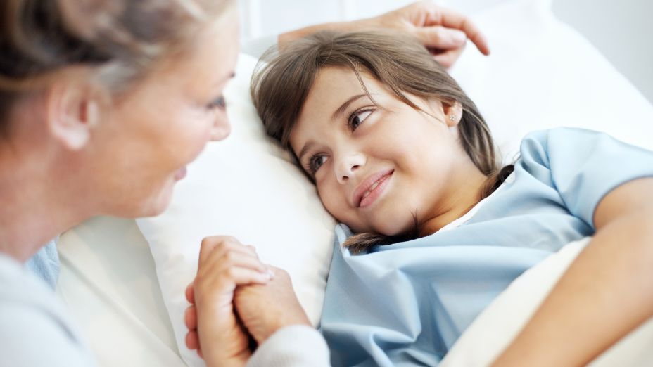 Ein Kind liegt im Bett und hat blaue Krankenhauskleidung an. Eine Frau ist vor dem Bett und hält die Hand des Kindes. Beide schauen sich in die Augen.