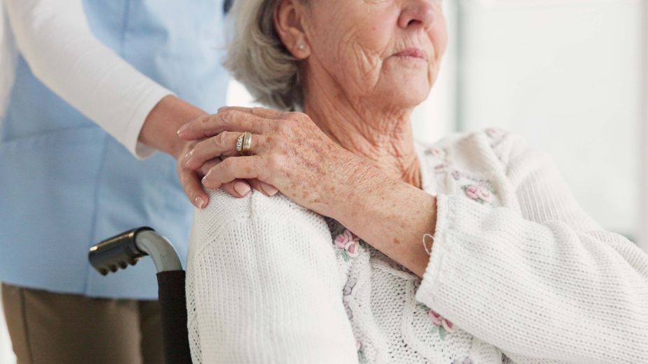 Eine Pflegerin steht hinter einer Frau im Rollstuhl und hat ihr die Hand auf die Schulter gelegt. Die Frau im Rollstuhl hat ihre Hand darauf gelegt