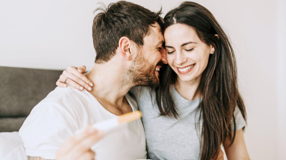 Eine Frau und ein Mann sind zu sehen. Beide haben die Köpfe aneinander gelehnt. Der Mann hält einen Schwangerschaftstest in der Hand. Beide lachen.