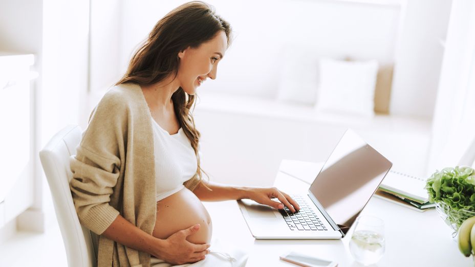 Eine schwangere Frau sitzt am Schreibtisch und schreibt mit ihrer linken Hand auf einem Laptop. Mit der rechten Hand hält sie ihren Babybauch.