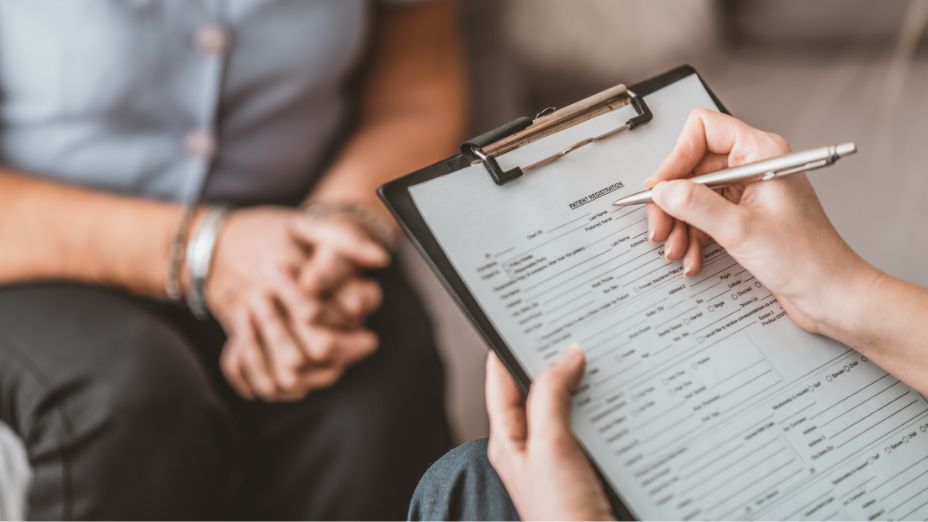 Im Vordergrund hält eine Person ein Klemmbrett in der Hand, auf welchem ein Blatt zu sehen ist. Im Hintergrund sitzt eine Person. 