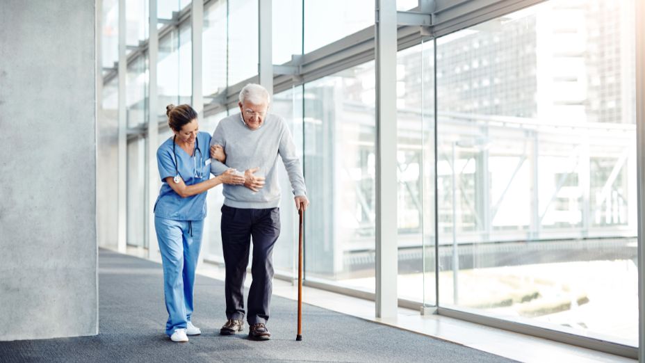 Eine Pflegerin geht neben einem älteren Herr und stützt ihn. Der Mann geht mit einem Gehstock.
