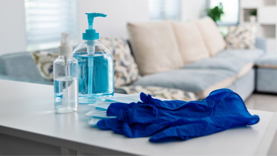Im Vordergrund liegen auf einem Tisch blaue Einweghandschuhe, eine Sprühfalsche mit durchsichtiger Flüssigkeit und eine Pumpflasche mit blauem Desinfektionsmittel.