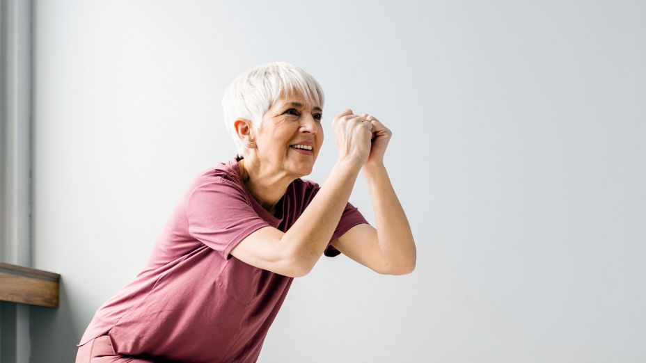 Eine Frau steht gebeugt vor einer weißen Wand und hält ihre Arme vor dem Körper zusammen. Die Hände sind zu Fäusten geformt und die Unterarme gehen dabei auseinander.