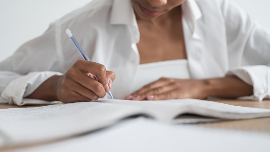 Eine Frau sitzt an einem Tisch und hat einen Stift in der Hand. Sie hat Hefte und Blätter vor sich liegen und schreibt dabei.