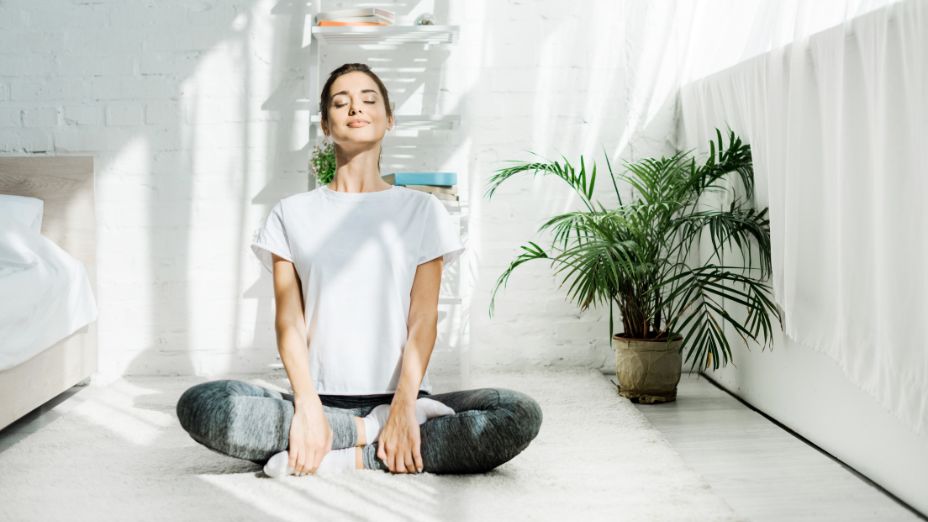 Eine Frau macht einen Schneidersitz und sitzt auf dem Boden. Dabei schaut sie zur Decke des Zimmers.