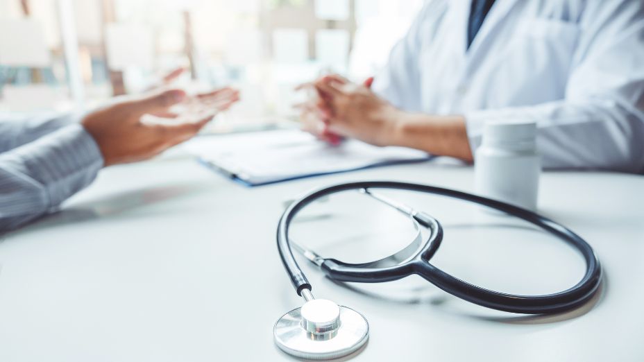 Im Vordergrund ist ein Stethoskop zu sehen. Dahinter verschwommen sind zwei Personen abgebildet. Diese sitzen gegenüber voneinander. Der eine hat einen Arztkittel an.