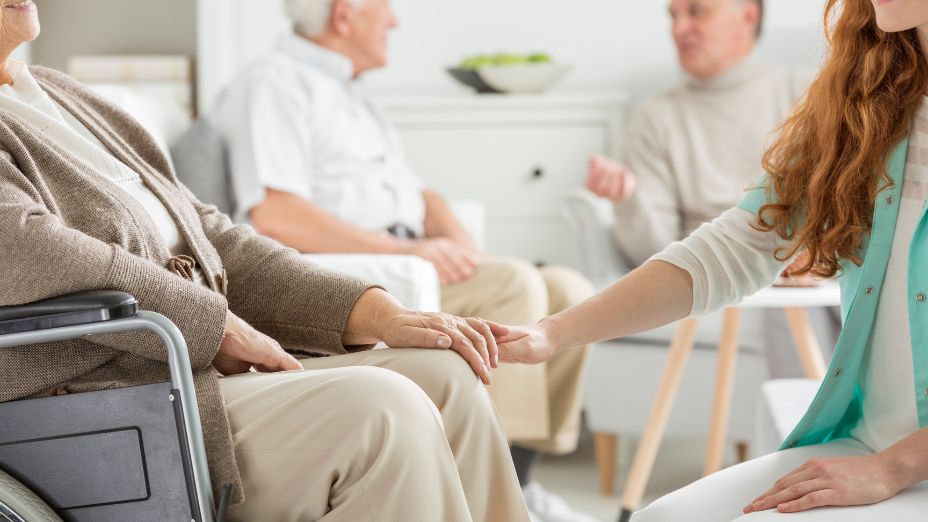 Im Hintergrund sitzen zwei Männer und reden miteinander. Im Vordergrund sitzt auf der linken Seite eine Frau im Rollstuhl und streckt ihre linke Hand aus. Auf der rechten Seite des Bildes sitzt im Vordergrund eine weitere Frau und streckt ihre rechte Hand aus. Beide Hände der Frauen liegen ineinander.