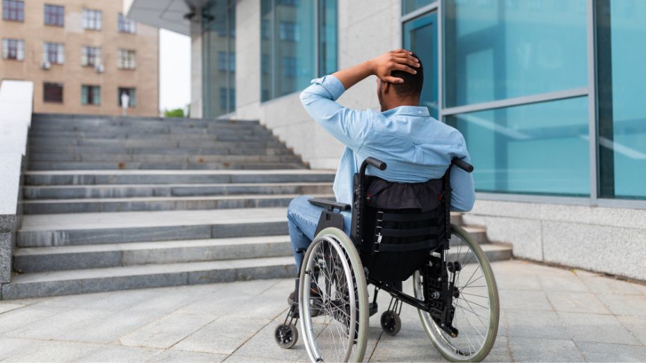Ein Mann sitzt im Rollstuhl und ist von hinten zu sehen. Mit seiner linken Hand fasst er sich an den Kopf. Er steht mit dem Rollstuhl vor einer Treppe und kommt nicht weiter.