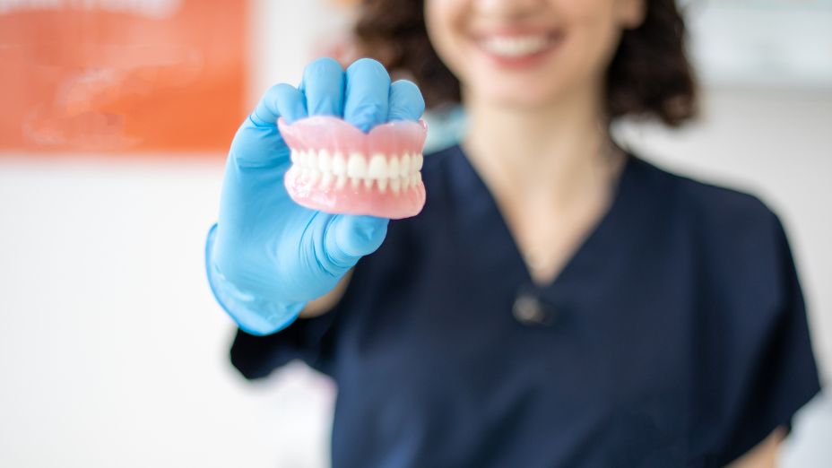 Eine Frau hält ein Gebiss in der Hand. Dieses ist scharf. Die frau im Hintergrund ist verschwommen.