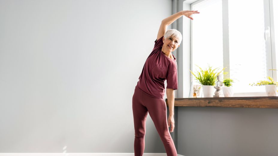 Eine ältere Frau macht Sport. Dabei steht sie mit den Beinen auseinander und hält eine Hand nach oben.